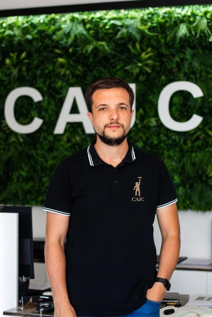 Um homem com uma camisola polo preta com um logótipo está em frente a uma parede verde exuberante, com as letras «CAJC» em destaque.