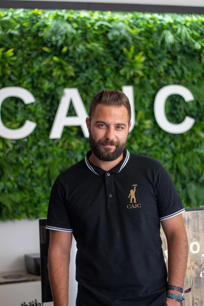 Um homem com barba e bigode está confiante em frente a uma parede verde com as letras «CAJC» parcialmente visíveis. Ele está a usar uma camisa polo preta com um logótipo dourado, transmitindo uma atmosfera profissional, mas descontraída.