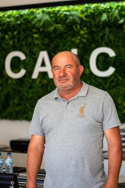 Um homem com uma camisa polo cinzenta está em frente a uma parede verde com as letras «CAJC» em branco, mostrando um ambiente moderno e interior.
