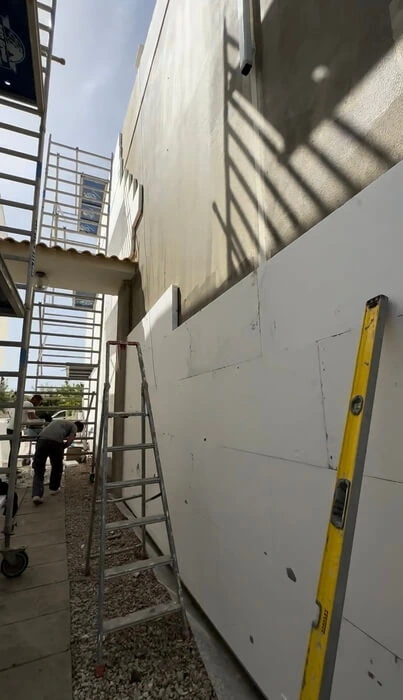 Um canteiro de obras com escadas, andaimes, um nível amarelo e painéis de parede exteriores inacabados sob a luz do dia.