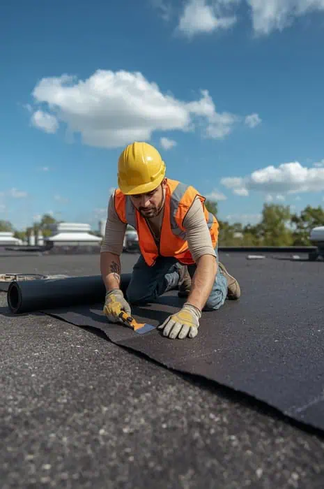 Um trabalhador da construção civil com capacete amarelo e colete de segurança instala material de cobertura num dia ensolarado, rodeado por nuvens.