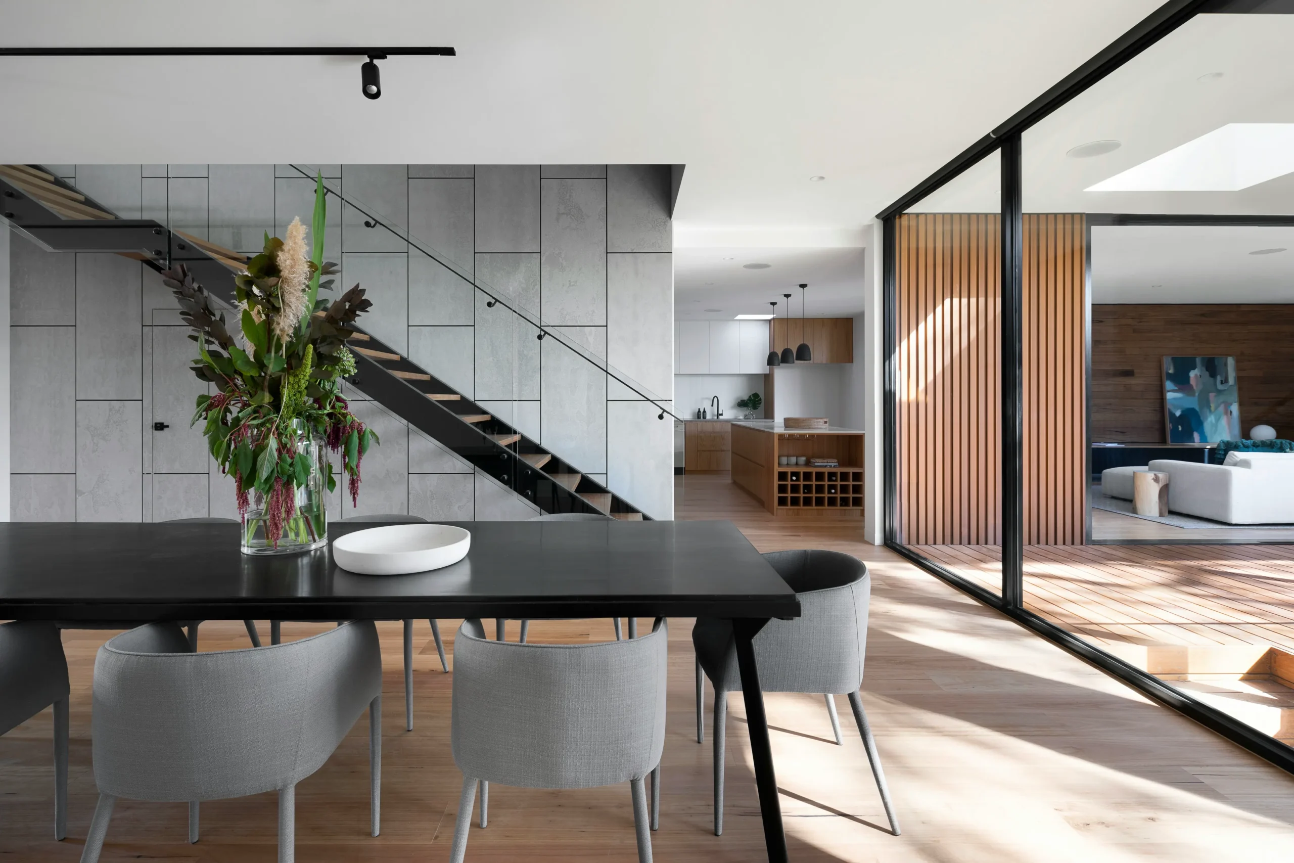 Design de interiores moderno com uma mesa de jantar preta e cadeiras estofadas cinzentas em primeiro plano, sobre um chão de madeira clara. Ao fundo, uma escada flutuante com corrimão preto contrasta com uma parede de painéis texturizados cinzentos. Vê-se uma cozinha aberta com armários claros e elementos em madeira, e uma porta de correr envidraçada preta que se abre para uma área de estar ou exterior com piso deck de madeira.