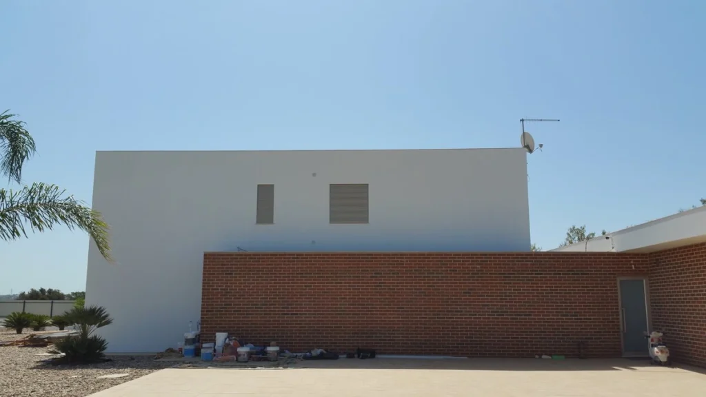 Fachada exterior de uma moradia moderna e minimalista sob céu azul. O design combina um volume branco retangular grande com duas pequenas aberturas, e uma parede de tijolo maciço vermelho em primeiro plano. O edifício está numa área exterior em construção ou em fase final de obra, com baldes de tinta e materiais no chão, e vegetação de paisagismo (palmeiras) à esquerda.