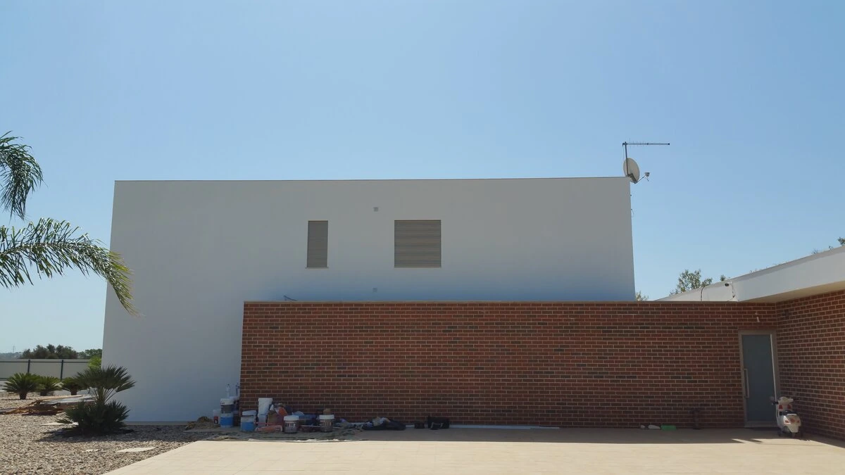 Fachada exterior de uma moradia moderna e minimalista sob céu azul. O design combina um volume branco retangular grande com duas pequenas aberturas, e uma parede de tijolo maciço vermelho em primeiro plano. O edifício está numa área exterior em construção ou em fase final de obra, com baldes de tinta e materiais no chão, e vegetação de paisagismo (palmeiras) à esquerda.