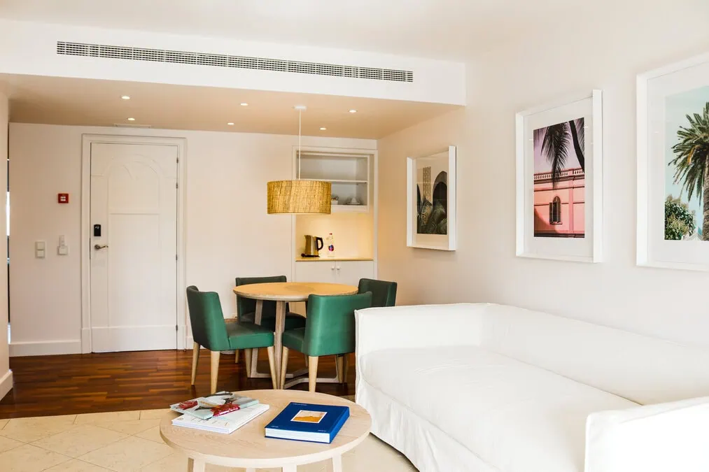 Interior de apartamento ou suite de hotel luminosa com decoração moderna e cores vibrantes. Em primeiro plano, um sofá branco e uma mesa de centro redonda clara. Ao fundo, uma mesa de jantar redonda de madeira com quatro cadeiras estofadas em veludo verde-esmeralda. As paredes brancas são decoradas com molduras brancas grandes com temas de palmeiras e arquitetura.