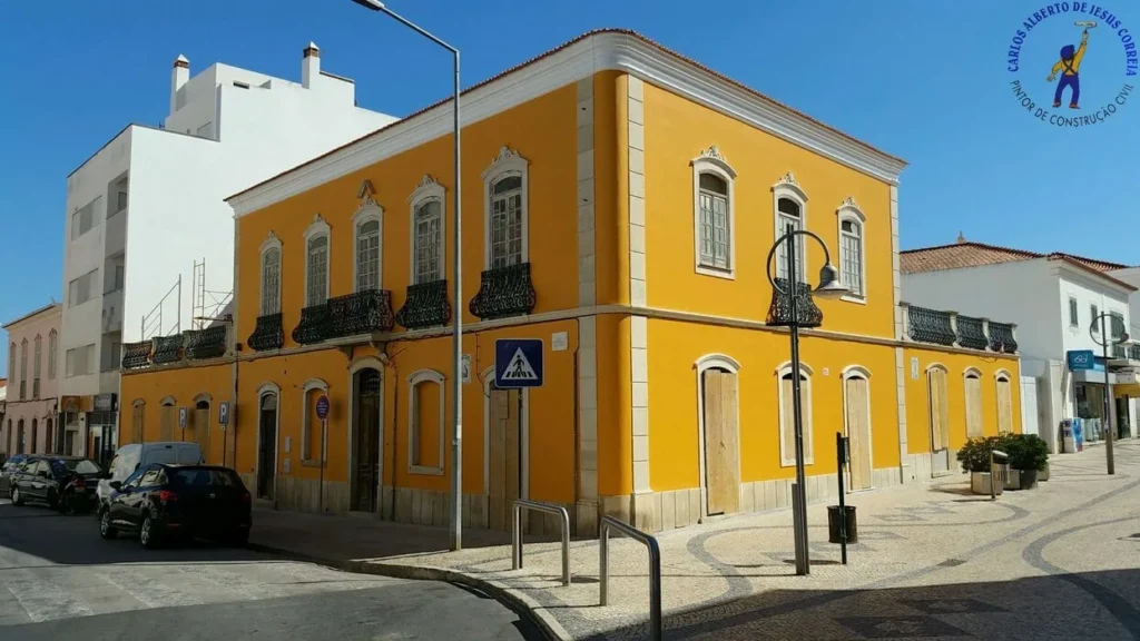 Fachada de um edifício de arquitetura tradicional portuguesa de dois andares, pintado de um amarelo mostarda vibrante com caixilharia branca, no Algarve (sugerido pelo estilo). O edifício tem sacadas de ferro forjado preto no primeiro andar e encontra-se num largo urbano com calçada portuguesa clara, rodeado por outros edifícios brancos.