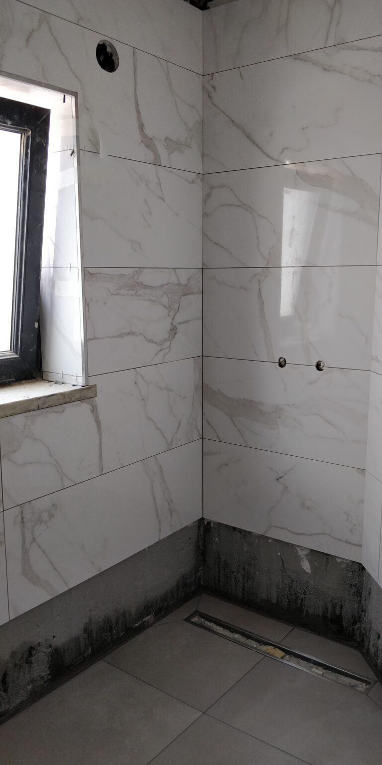Divisão interior (casa de banho ou duche) em fase de construção com revestimento de paredes em azulejos de grande formato com efeito mármore Calacatta ou Carrara (branco com veios cinzentos). A imagem mostra o canto da divisão e o nicho da janela com caixilharia escura, e a base de duche ou pavimento em cimento/cimento queimado cinzento, com o escoamento linear já instalado. A fase de acabamentos está em curso.