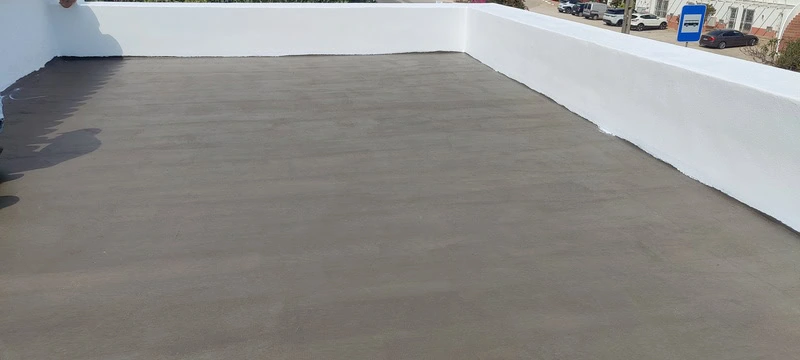 Uma visão ampla de um terraço já impermeabilizado, com a superfície coberta por uma camada uniforme e cinzenta. Um claraboia retangular branca é visível no lado direito. O contraste entre o novo revestimento e as paredes brancas realça a qualidade do trabalho.