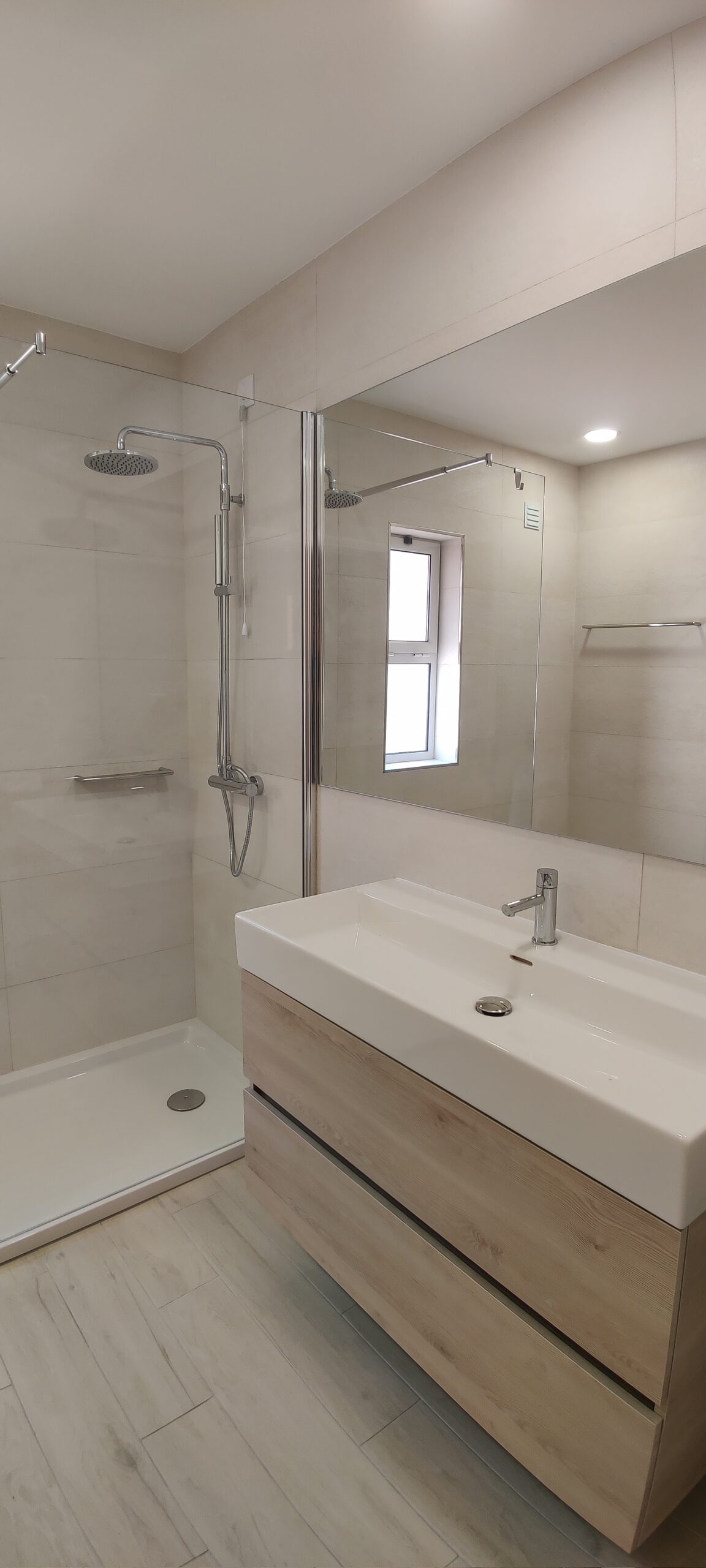 Casa de banho moderna e minimalista com cores neutras. Destaque para o lavatório de pousar em cima de um móvel suspenso de madeira clara com gavetas, um espelho grande que duplica o espaço e uma base de duche walk-in com resguardo de vidro. O revestimento em azulejo bege claro e o pavimento em cerâmica ou vinílico com padrão de madeira clara conferem um ambiente clean e acolhedor.
