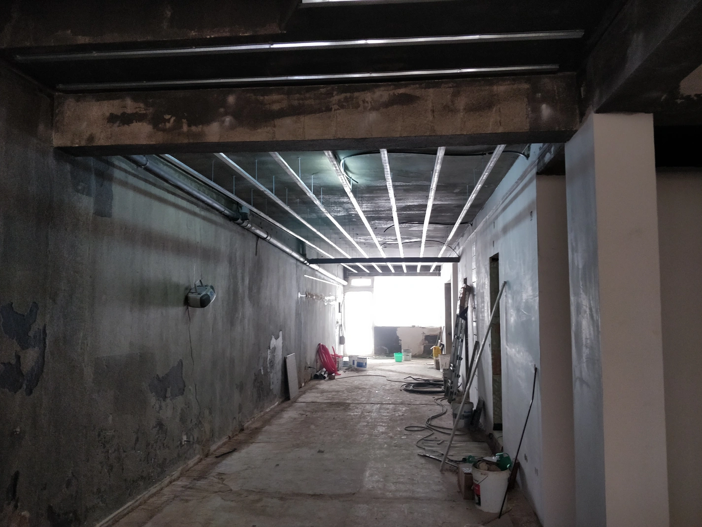 Corredor em construção com teto em laje de betão e vigas, onde se instala sistema de teto falso com calhas metálicas. Remodelação de espaço comercial/industrial, paredes brutas.