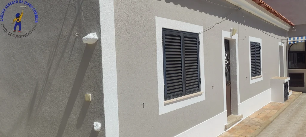 Detalhe de janela com persiana preta numa fachada de casa reabilitada. A pintura exterior em cinzento claro e a moldura branca realçam o acabamento texturado. Demonstração de trabalho de pintor de construção civil em Portugal.